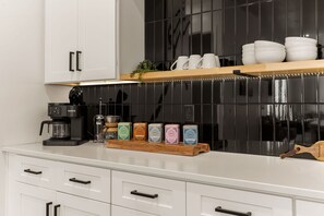 A kitchen with white cabinets, a black tiled backsplash, a coffee maker, and a shelf with cups and jars. - On The Rocks Shaken 6 Bedroom Home (Hurricane)