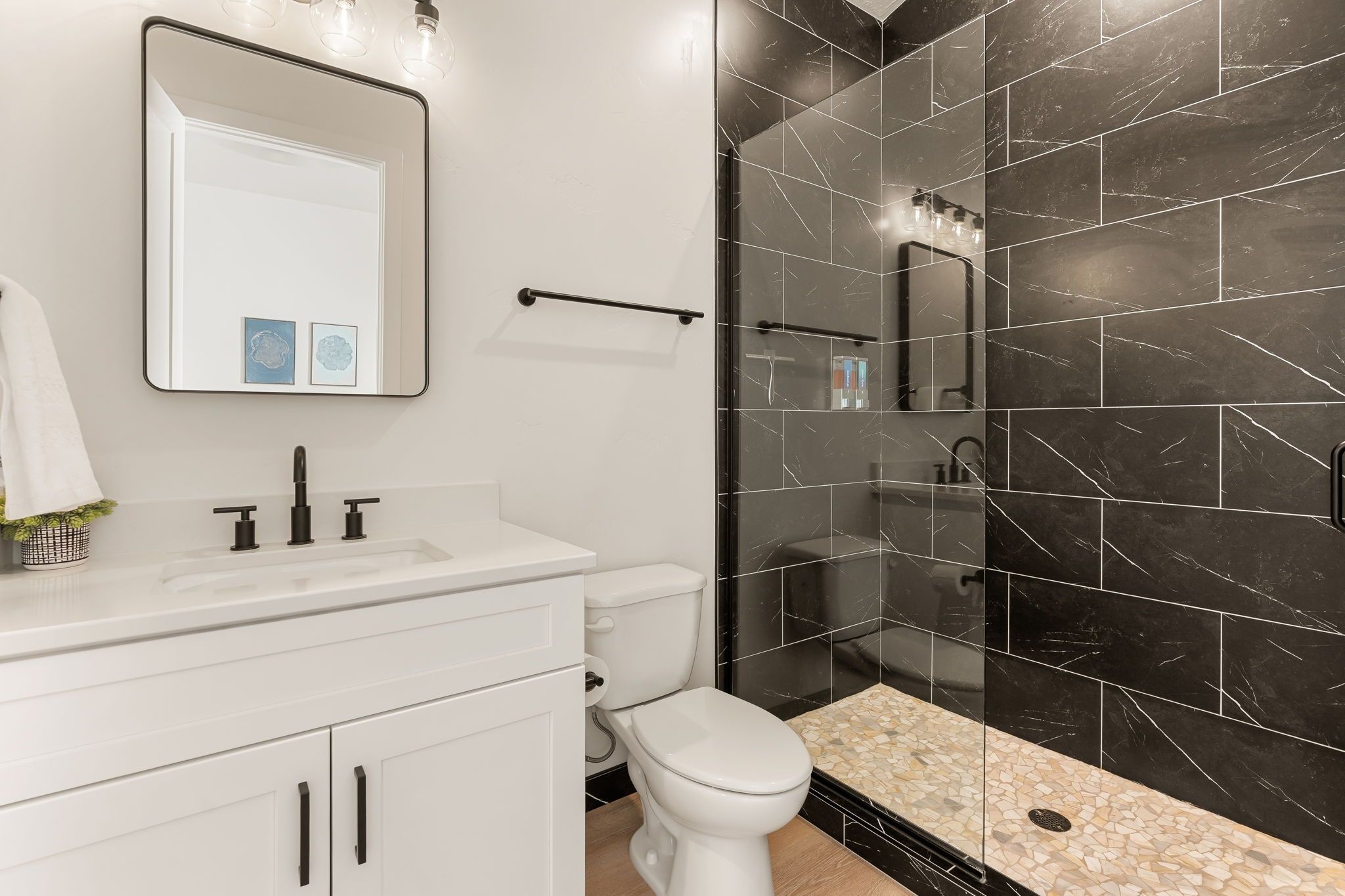 A modern bathroom with a white vanity, a toilet, a shower with black tiles, and a mirror with wall-mounted lights.