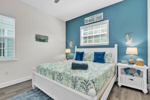 A bedroom with a bed, two bedside tables, a nightstand, a window with blinds, and a wall sign. - Blue Star Oasis 4 Bedroom Home (Holmes Beach)