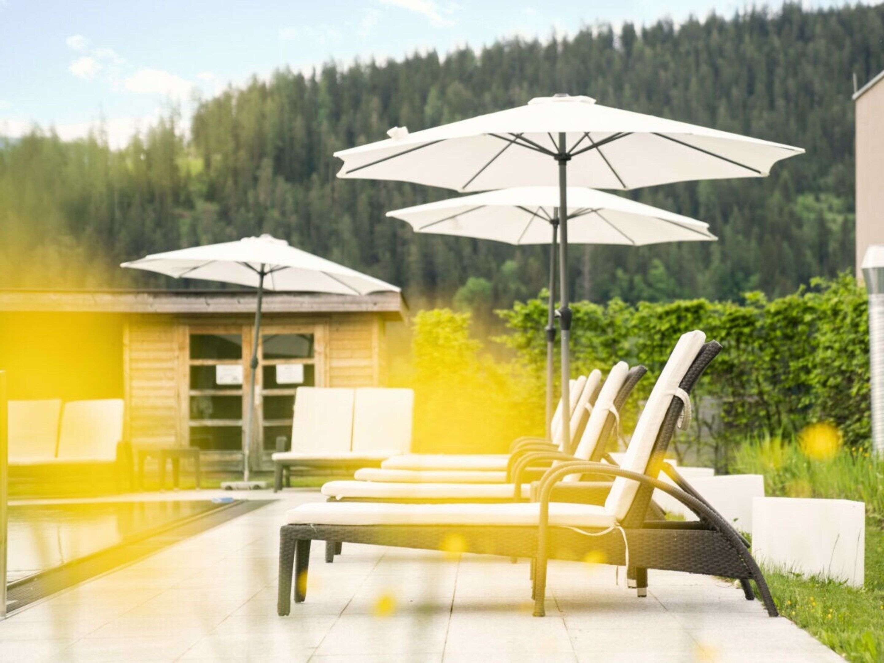 Ein Poolbereich mit Liegestühlen und Sonnenschirmen, im Hintergrund ein Holzgebäude und ein bewaldeter Hügel.