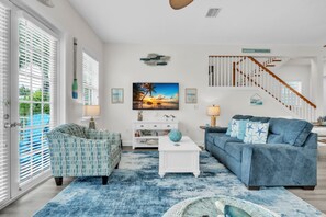 A spacious living room with a blue sofa, a patterned armchair, and a white coffee table. A large painting of a sunset is on the wall, and there is a staircase with wooden railings. - Blue Star Oasis by Duncan Real Estate (Holmes Beach)