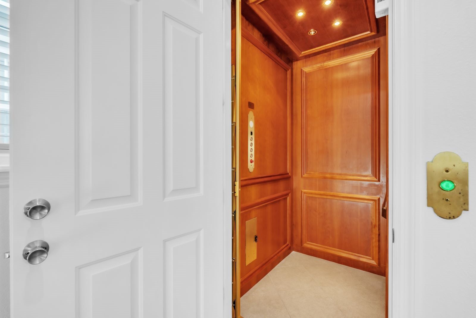 Puerta blanca con portero automático de latón y una luz verde indicadora, que da acceso a un ascensor de paneles de madera con un panel de control visible.