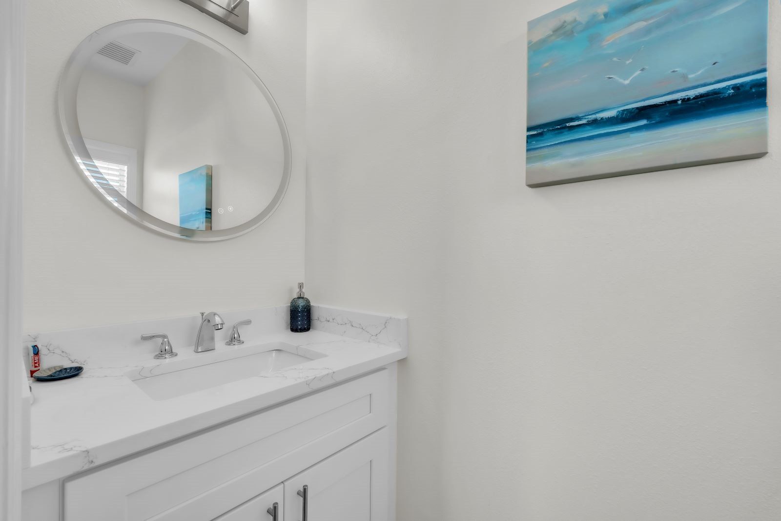 A bathroom with a white vanity, a round mirror, a sink, and a wall-mounted painting.