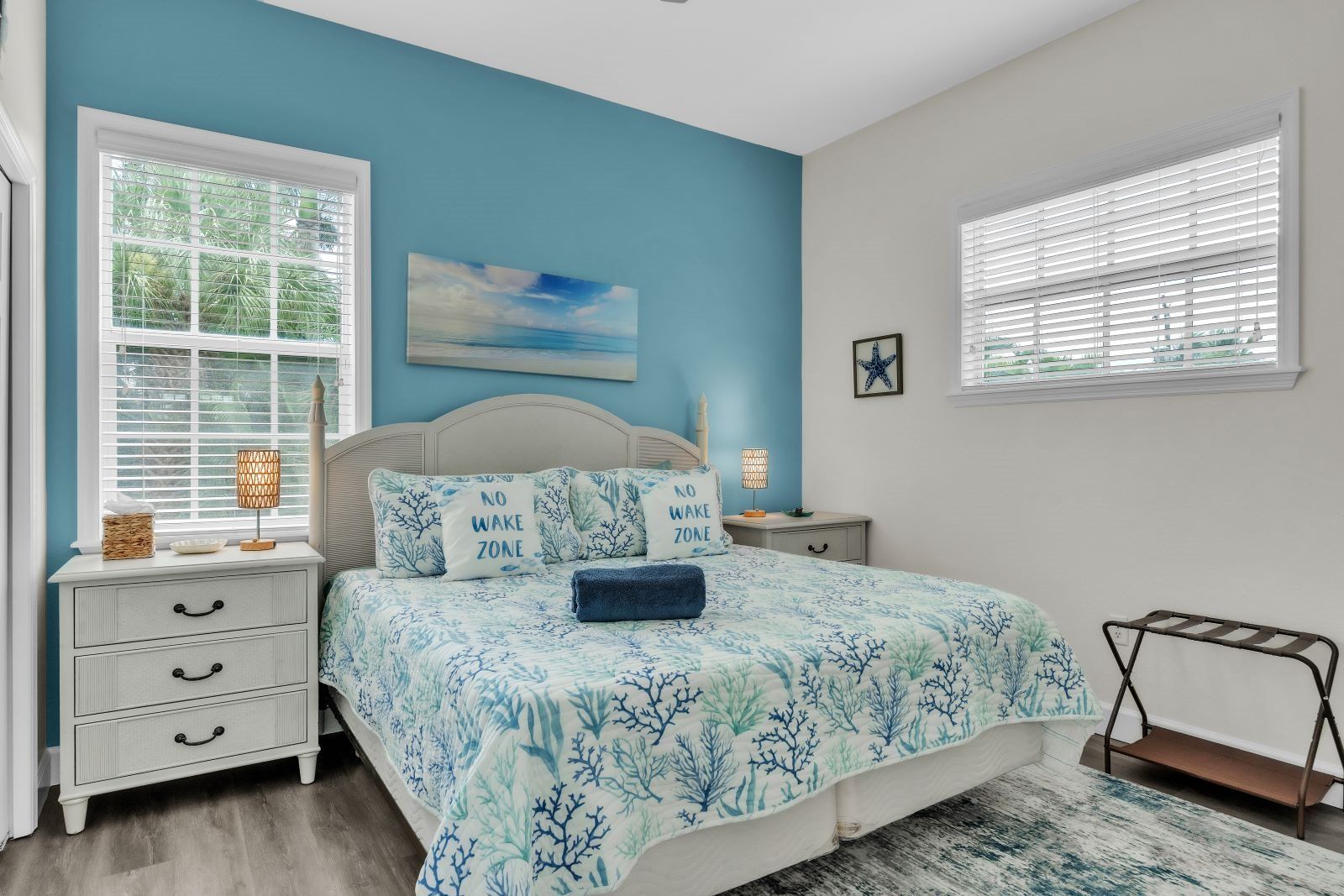 A bedroom with a bed, two bedside tables, a nightstand, a window with blinds, a starfish decoration, and a painting on the wall.