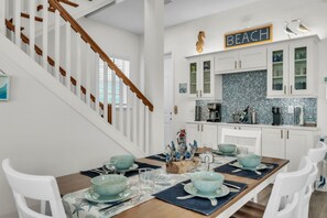Un coin repas avec une table en bois dressée pour quatre personnes, des chaises blanches et une cuisine dotée de meubles blancs et d’un dosseret de cuisine en mosaïque bleue.