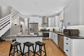 Una cucina moderna con mobili bianchi, piano cottura in acciaio inossidabile, forno a microonde e forno integrato. Sono presenti tre sgabelli neri all'isola.