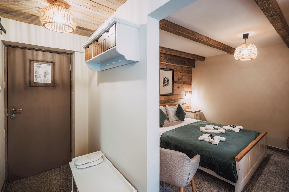 Habitación de hotel con techo de madera, cama con colcha verde, mesita de noche y lámpara colgante.
