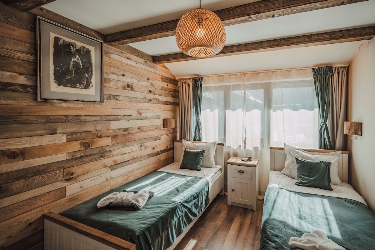 Una habitación con paneles de madera, dos camas con ropa de cama verde, una mesita de noche, una lámpara colgante y una ventana con cortinas.