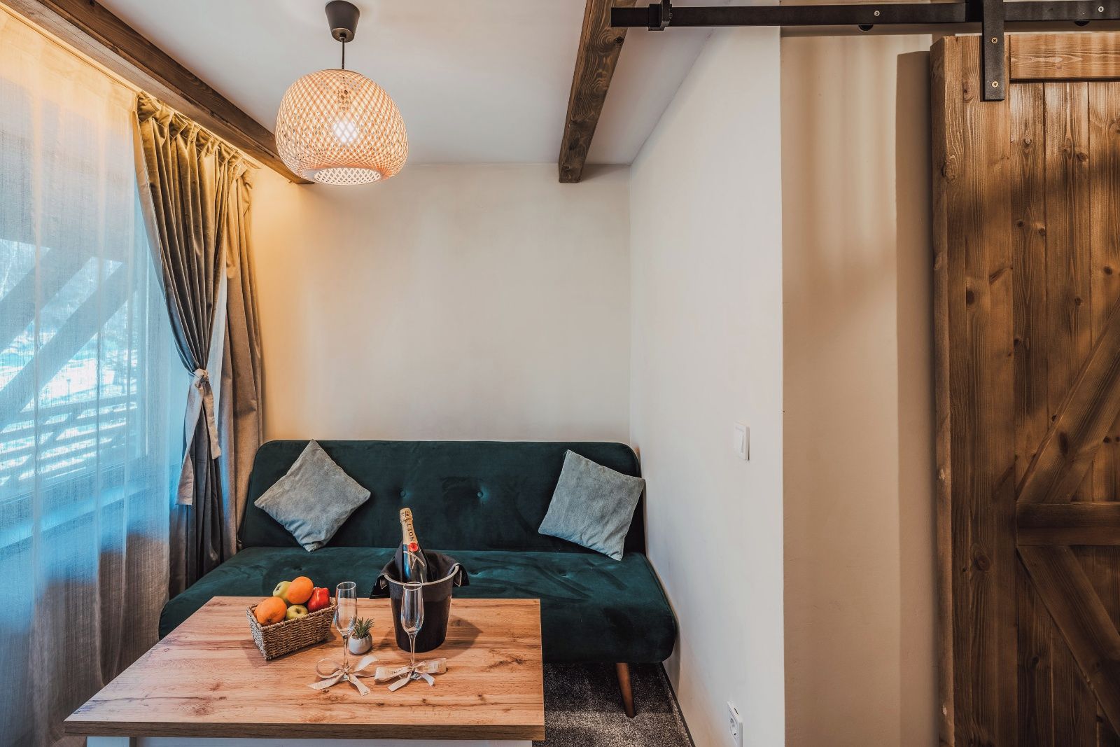 A cozy room with a green sofa, a wooden table with a bottle and glasses, and a hanging light fixture.