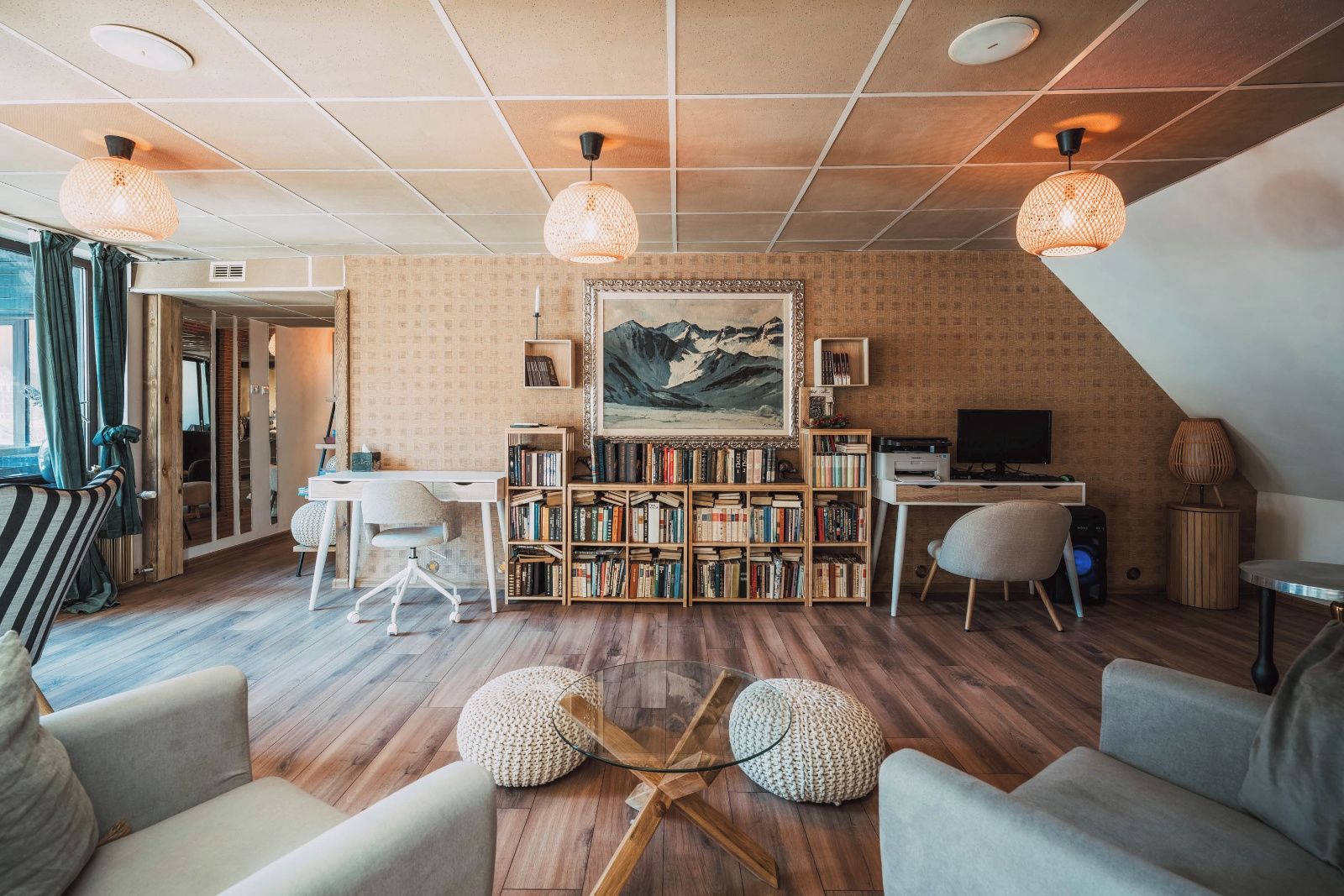 A cozy living room with a wooden floor, a bookshelf, a desk with a computer, and a large painting on the wall.