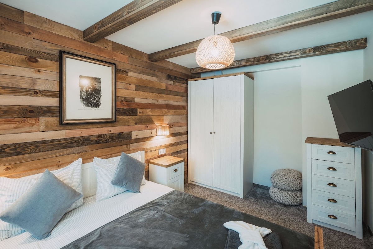Un dormitorio con pared de madera, una cama con almohadas, una mesita de noche con lámpara, un armario y un televisor sobre un aparador.