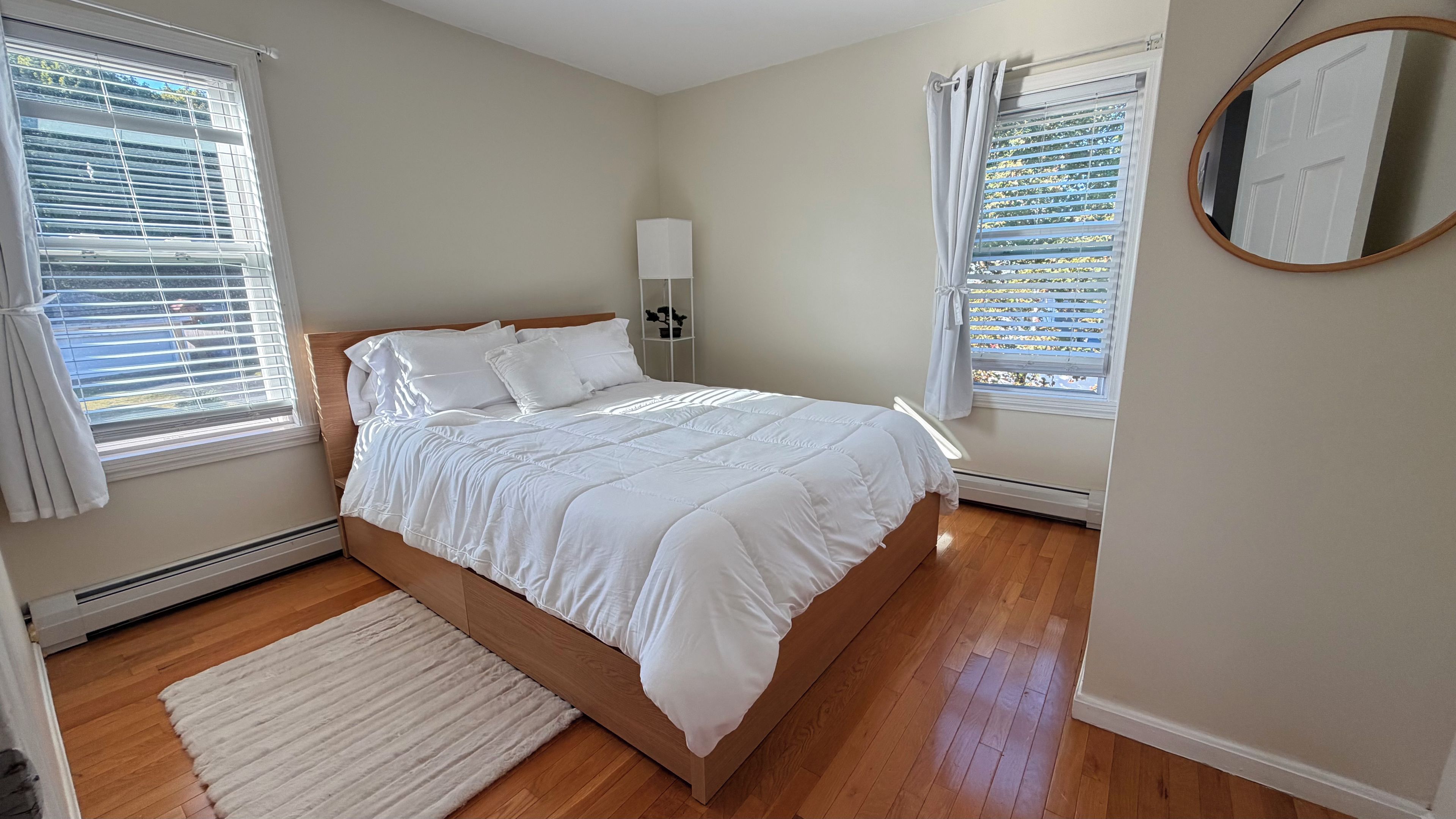 A bedroom with a bed, two windows with blinds, a bedside table with a lamp, and a mirror on the wall.