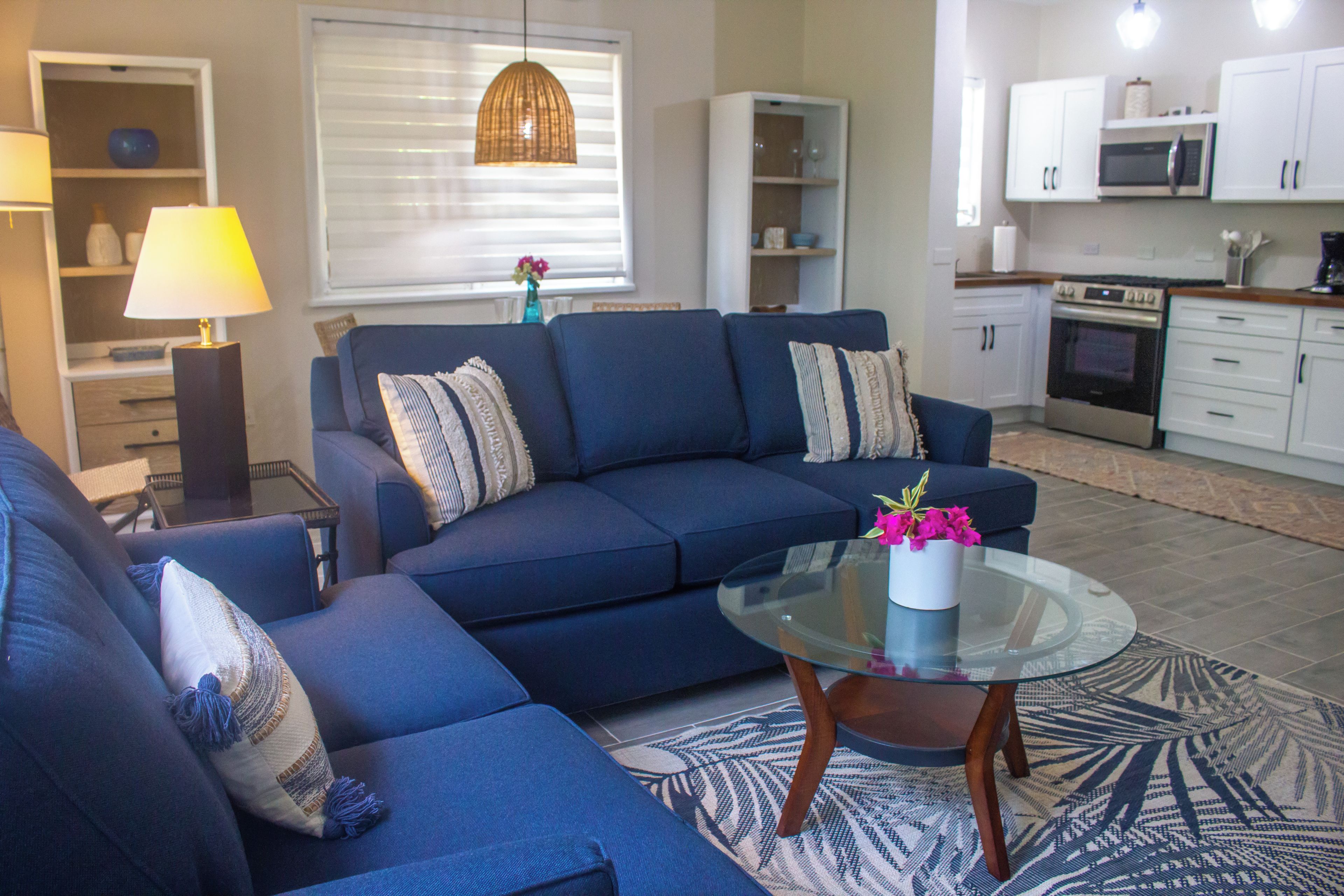 A living room with a blue sofa set, a glass coffee table with a vase of flowers, and a kitchen in the background.