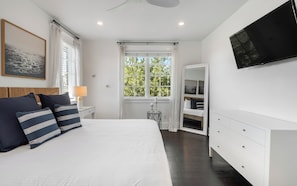Ein Schlafzimmer mit einem Bett, Nachttisch, Lampe, Fenster und einem an der Wand befestigten Fernseher.