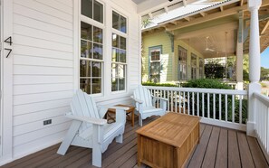 A wooden deck with two Adirondack chairs and a small table, overlooking a green house with white trim. - Salt and Pine | Golf Cart, Pool, Bikes (Santa Rosa Beach)