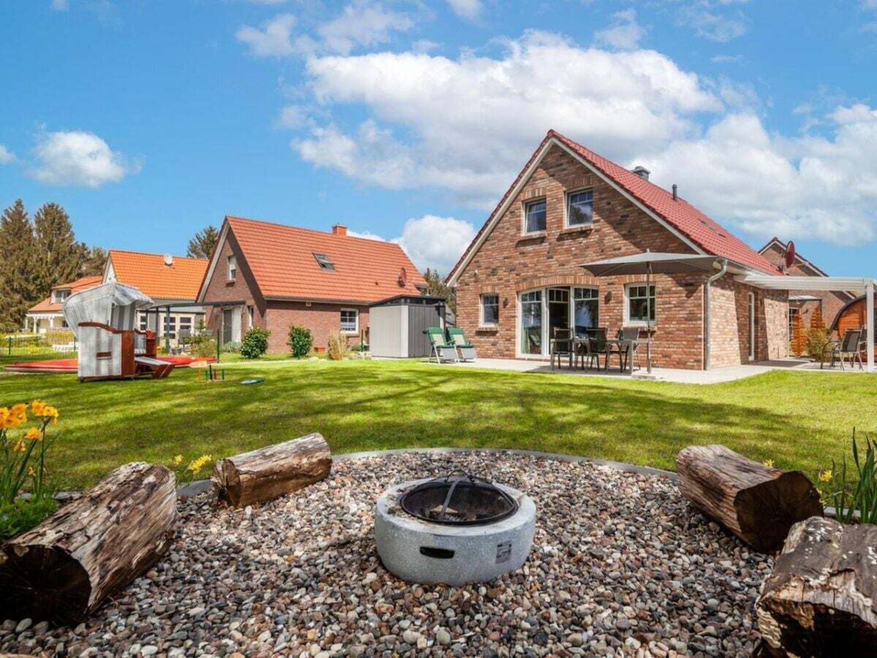 Une grande maison en briques avec un toit rouge, une terrasse couverte et un foyer entouré de bûches et de galets.