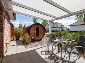 Ein überdachter Patio-Bereich mit einer holzverkleideten, tonnenförmigen Struktur, Gartenmöbeln und Blick auf Wohnhäuser im Hintergrund.