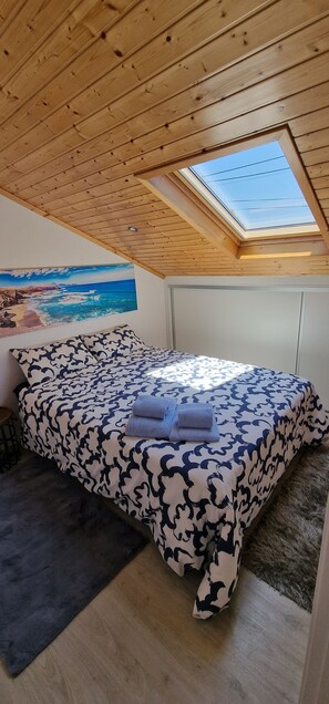 Quarto com teto de madeira, claraboia, cama com roupa de cama estampada e quadro com paisagem de praia.