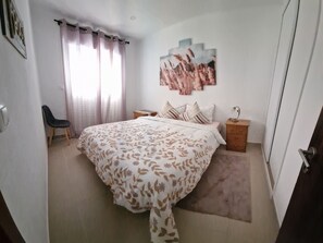A bedroom with a bed, a nightstand, a chair, a window with curtains, and a wall-mounted artwork. - Sunset House Meco pôr do Sol & Praia a 1500 m (Alfarim-sesimbra)
