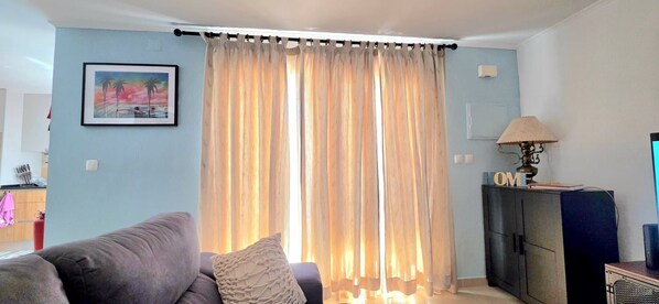Ein Wohnzimmer mit einer Couch, einer Lampe, einem Fernseher und einem Fenster mit Vorhängen.