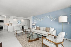 A modern living room with a glass coffee table, two armchairs, a sofa, and a kitchen in the background. - Sky-High Beachfront Paradise: Updated Condo with Resort Amenties. Walk to Dining & Shopping Hotspots (Marco Island)