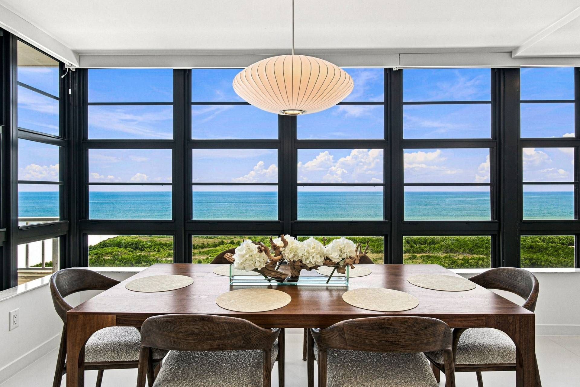 Un espace repas comprenant une grande table, des chaises, un vase de fleurs et une vue sur l’océan grâce à de grandes fenêtres.