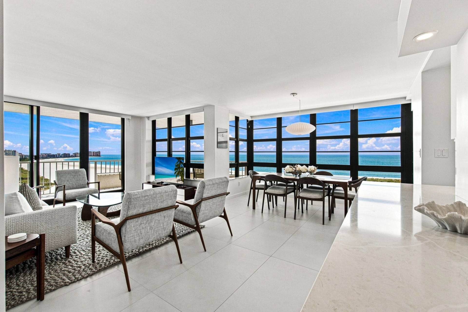 A spacious living area with a large window overlooking a beach, furnished with modern chairs and a dining table.