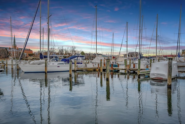 A marina with multiple boats docked at wooden piers, surrounded by buildings and trees.
