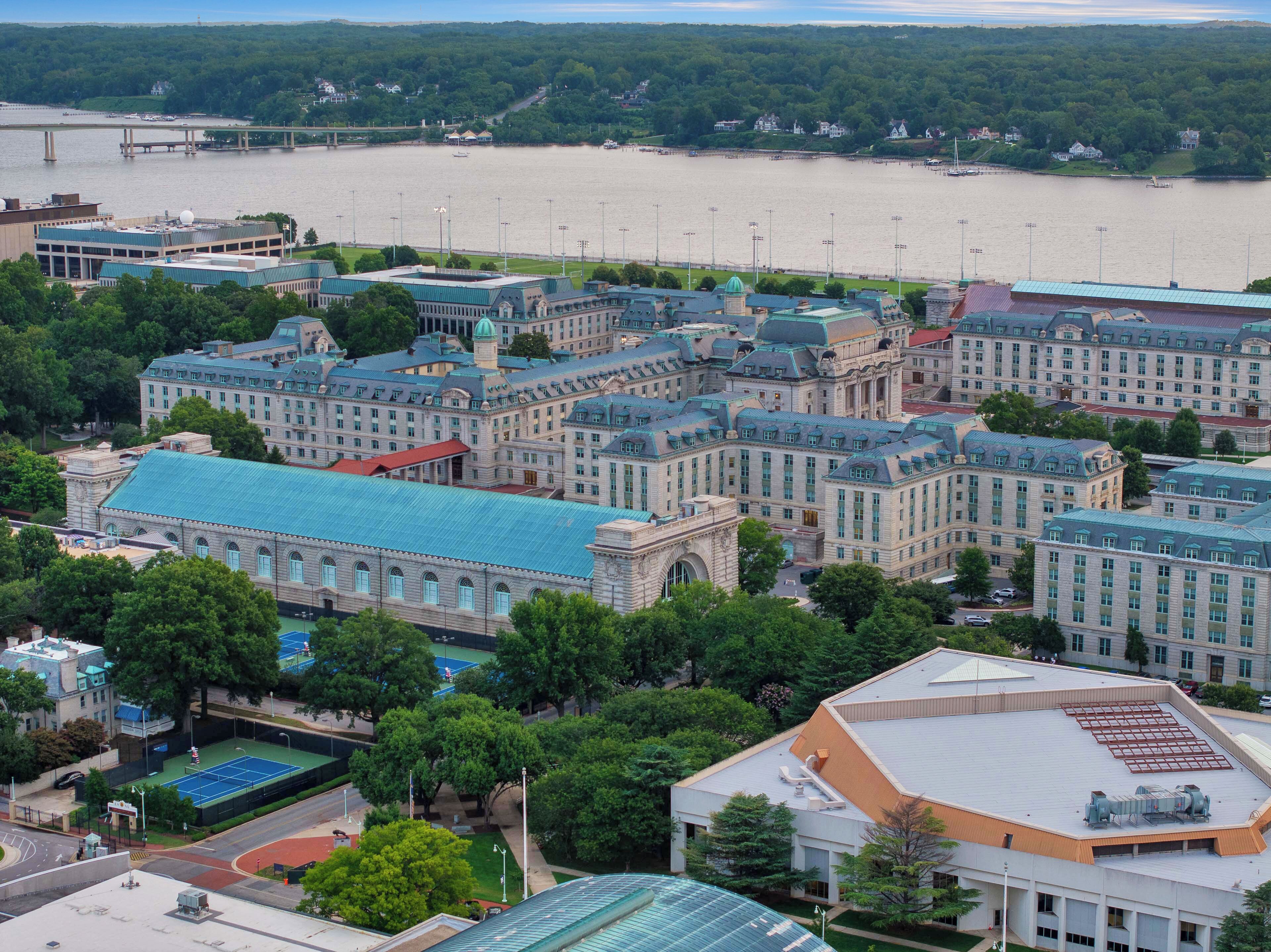 A large institutional building with multiple wings, a tennis court, and a river in the background.