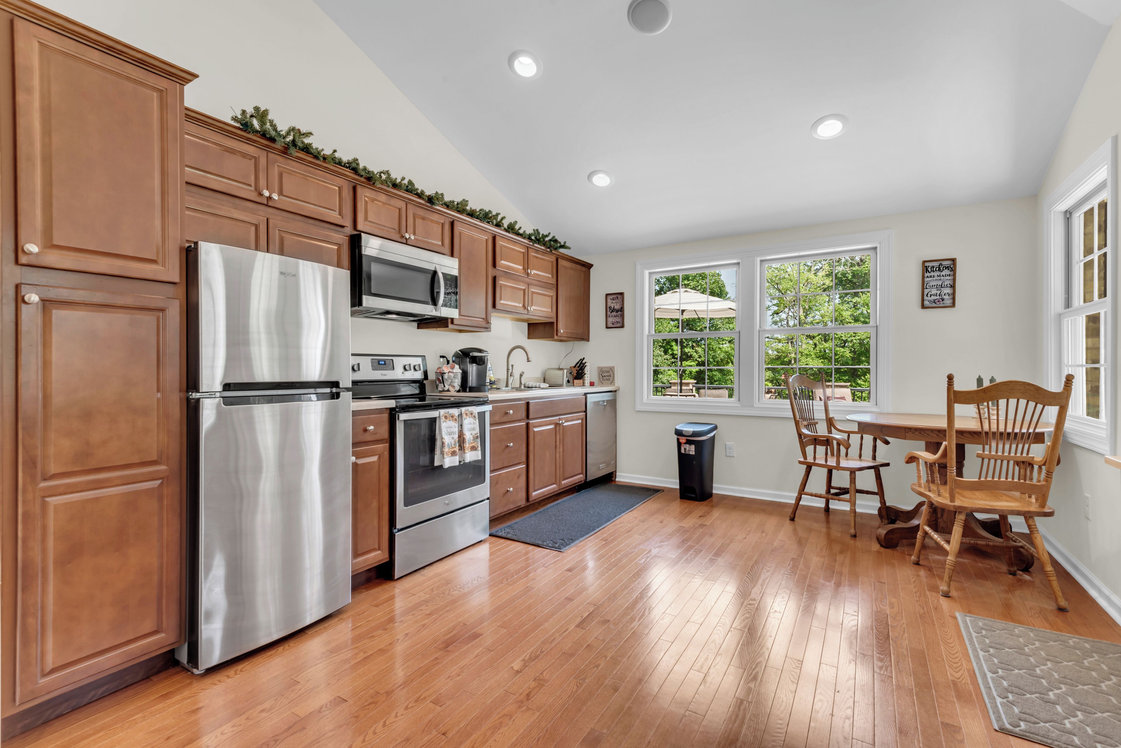 A kitchen with wooden cabinets, stainless steel appliances, a dining area with wooden chairs, and a view of greenery through the windows.