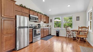 A kitchen with wooden cabinets, stainless steel appliances, a dining area with wooden chairs, and a view of greenery through the windows.