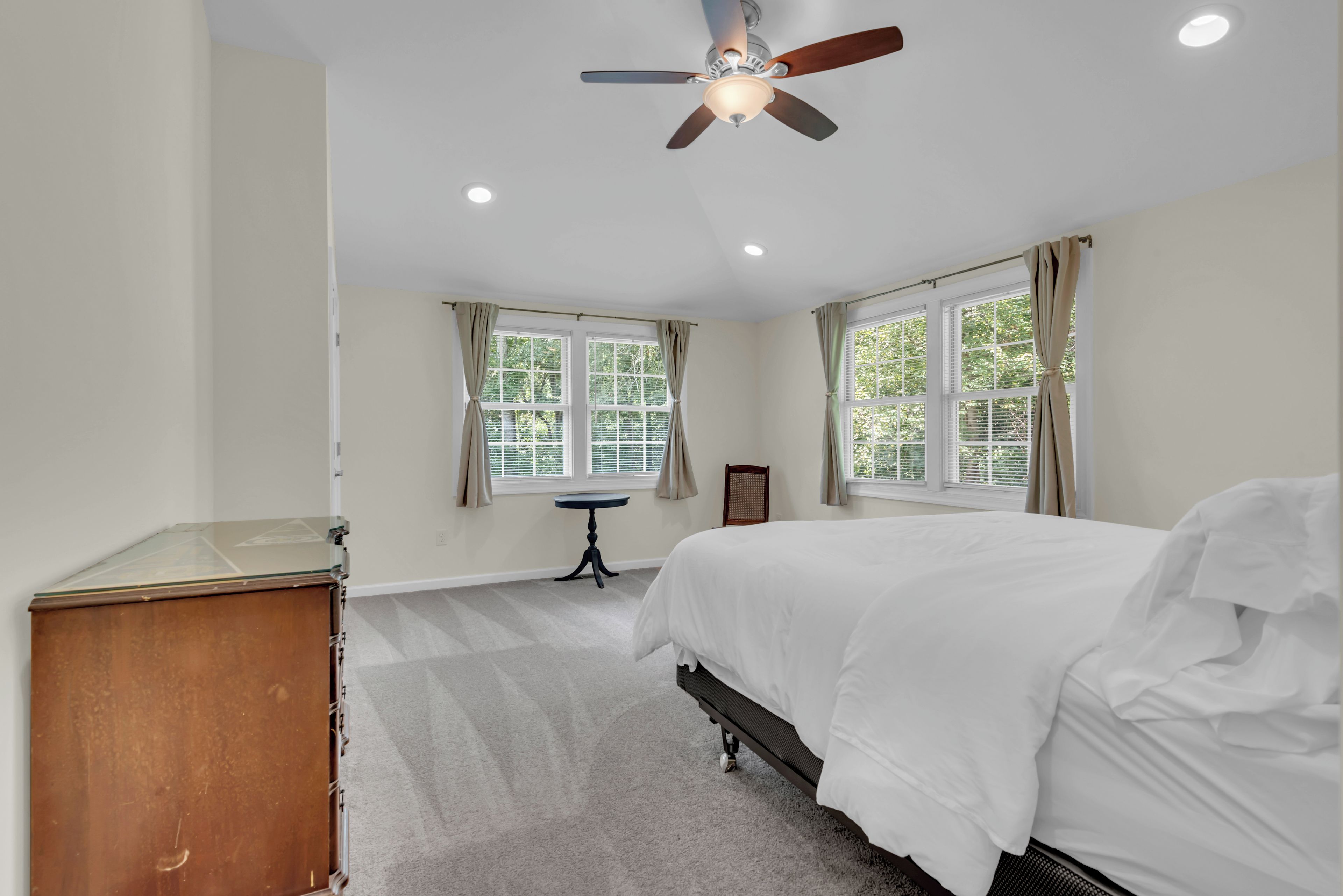Quarto com cama, ventoinha no teto, duas janelas com cortinas, uma mesa pequena e um armário.
