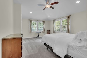 A bedroom with a bed, a ceiling fan, two windows with curtains, a small table, and a dresser.