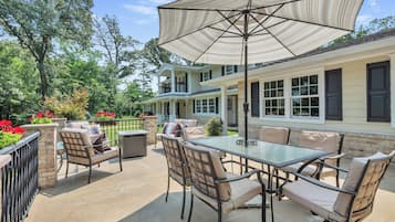 Une terrasse extérieure avec une table et des chaises en verre, un grand parasol, et une maison dotée de nombreuses fenêtres et d’un balcon.