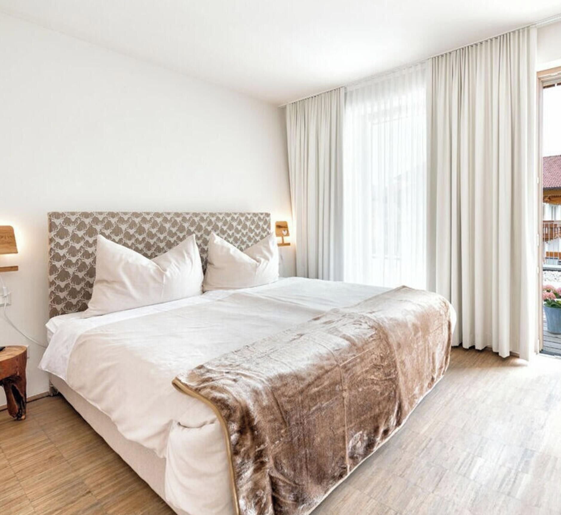 A hotel room with a large bed, a headboard with a patterned design, two bedside lamps, and a view of a balcony with curtains.