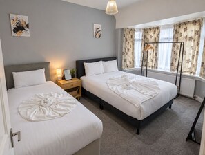 A hotel room with two beds, a bedside table with a lamp, a window with curtains, and artwork on the walls. - Spacious 4-bed Southall | Free Parking | Sleeps 9 (Southall)