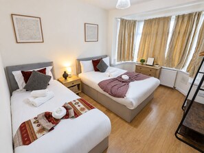 A hotel room with two beds, a bedside table with a lamp, a window with curtains, and a radiator. - Spacious 4-bed Southall | Free Parking | Sleeps 9 (Southall)