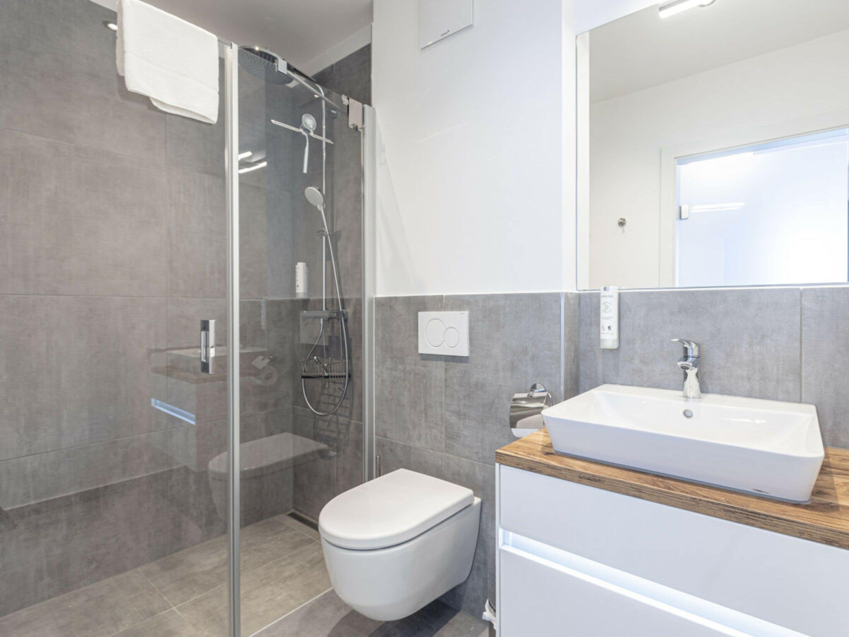 Un baño moderno con ducha envuelta en vidrio, inodoro blanco y tocador de madera con lavamanos.