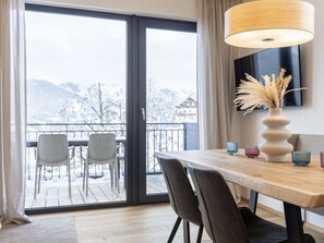 A dining area with a wooden table, chairs, a vase with pampas grass, and a view of a snowy mountain landscape through a glass door. - Apartment With 1 Bedroom (Sankt Martin am Tennengebirge)