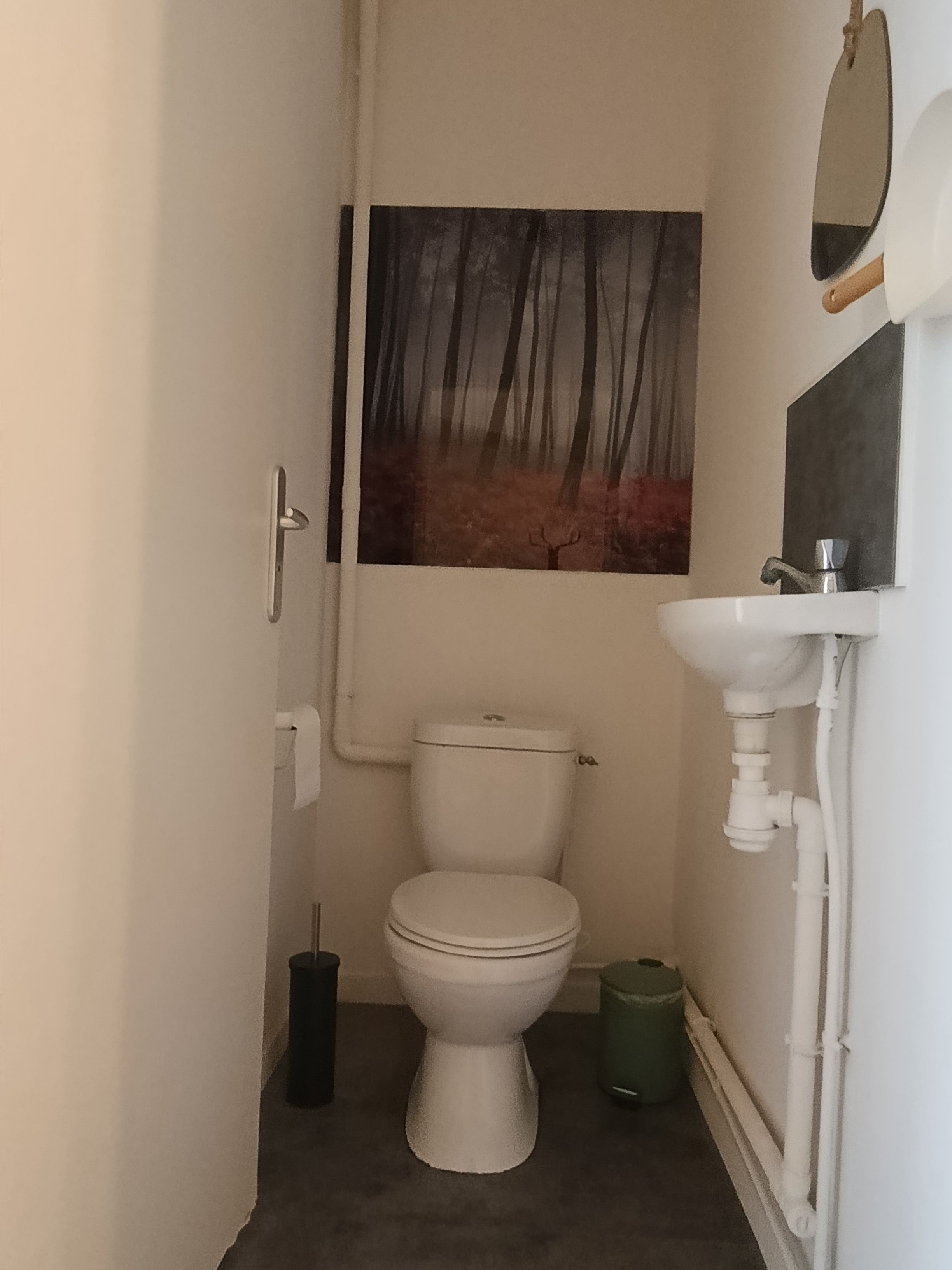 Ein kleines Badezimmer mit Toilette, Waschbecken, Spiegel und einem Waldgemälde an der Wand.
