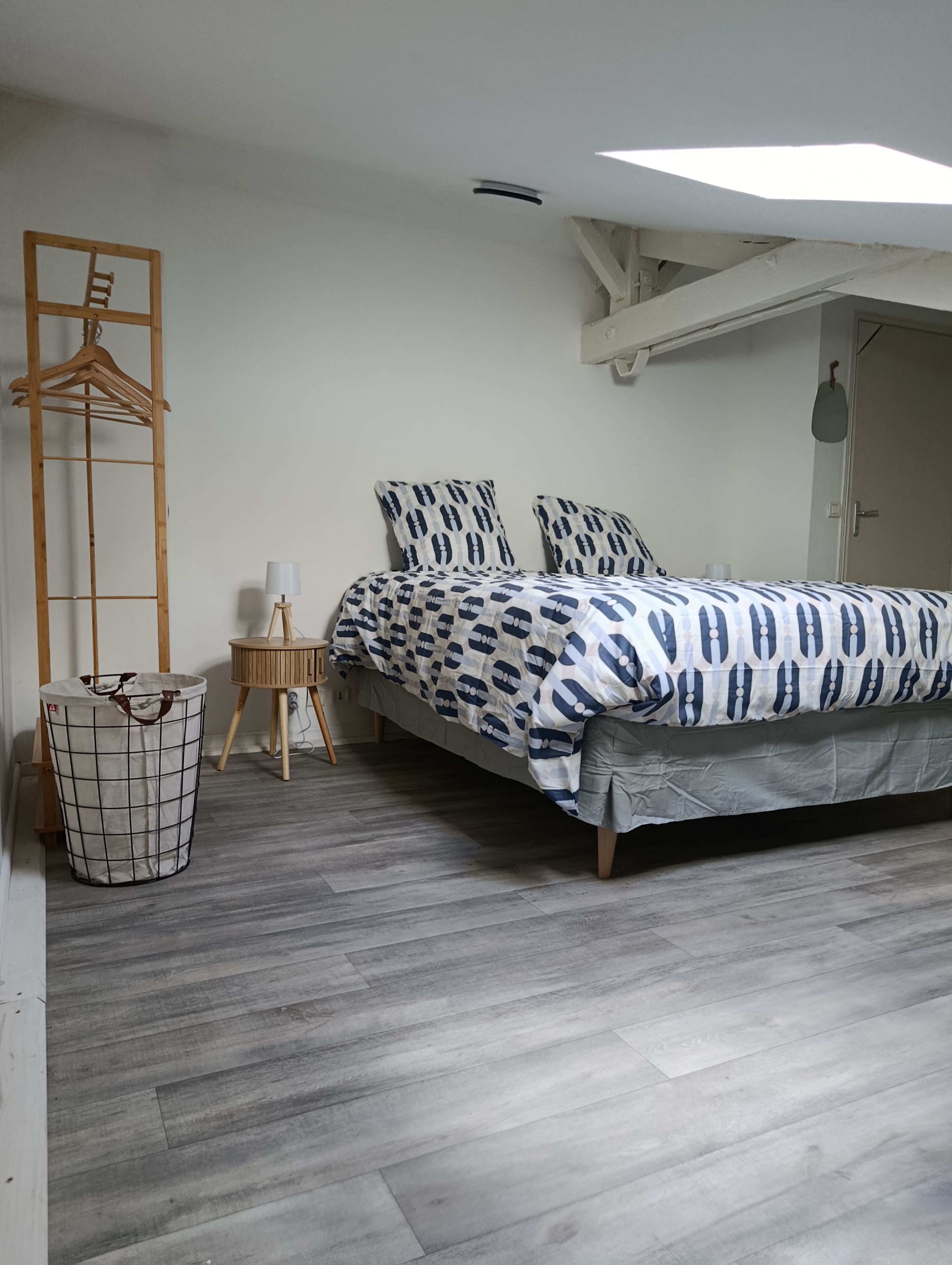 Ein Schlafzimmer mit einem Bett, einem Holzgalgen, einer Lampe, einem Mülleimer und einer Deckenleuchte.