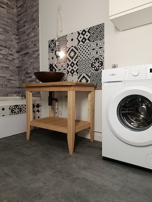 A laundry area with a washing machine, a wooden table with a bowl, and patterned tiles on the wall.