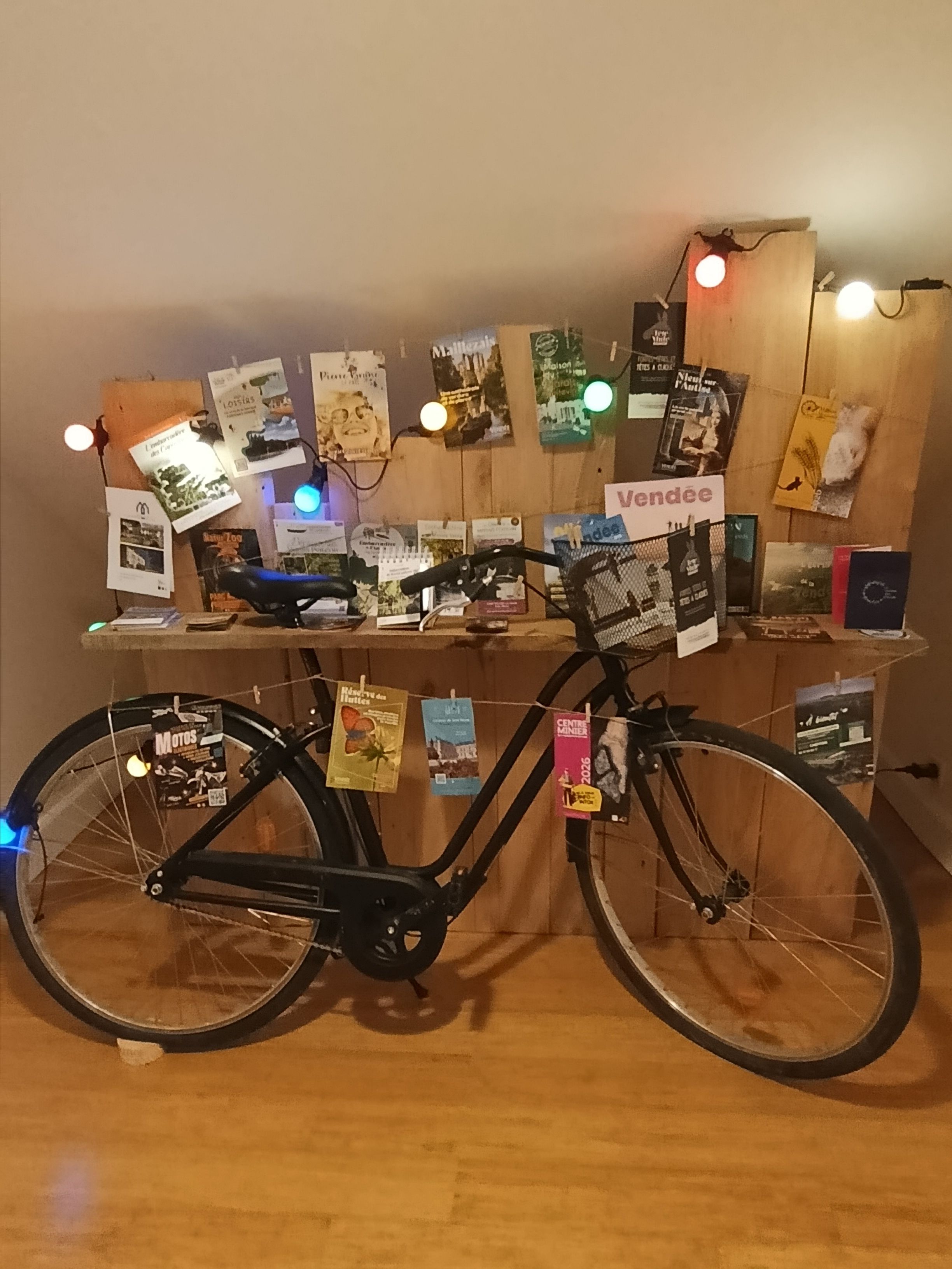 Una bicicletta con un cesto, una scrivania di legno con diversi oggetti e documenti, e una parete con lampadine e poster.
