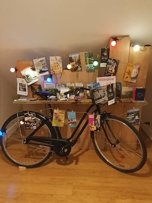 A bicycle with a basket, a wooden desk with various items and papers, and a wall with light bulbs and posters.