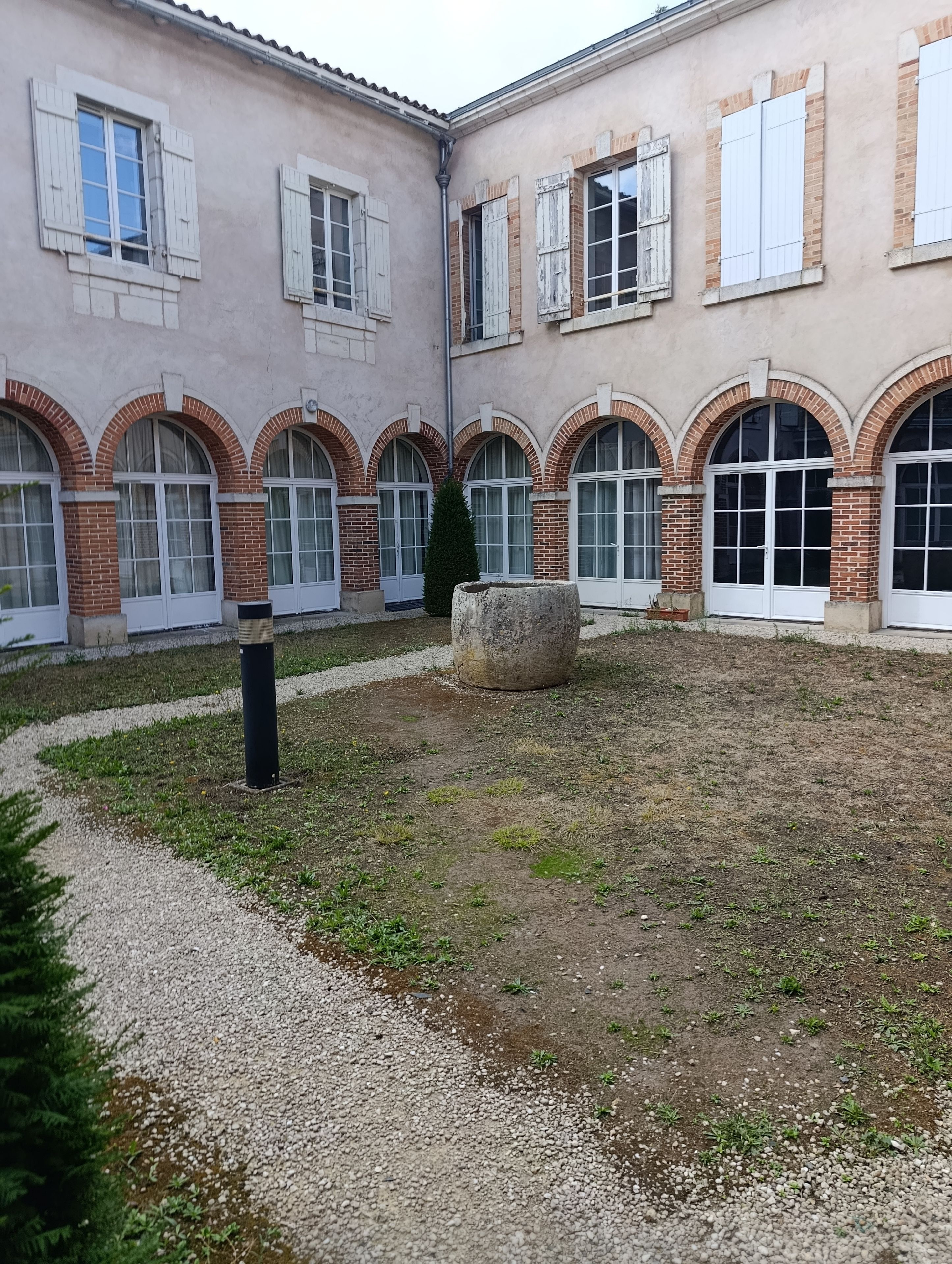 Un cortile con un vialetto sterrato, un vaso in pietra e un edificio con finestre ad arco e mattoni a vista.