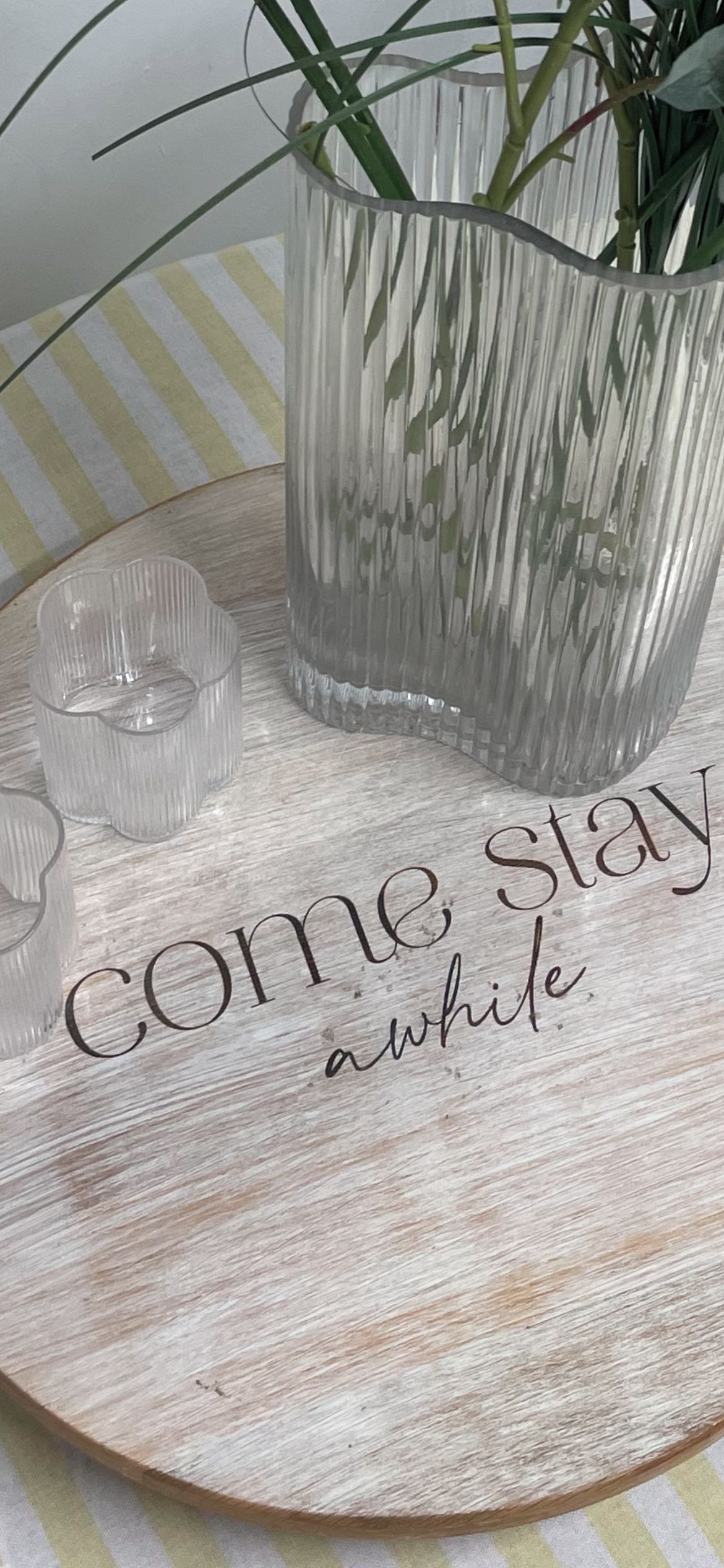 Ett träbricka med en vas i glas och en ljusstake, samt en träskylt med texten ”stanna till en stund”.