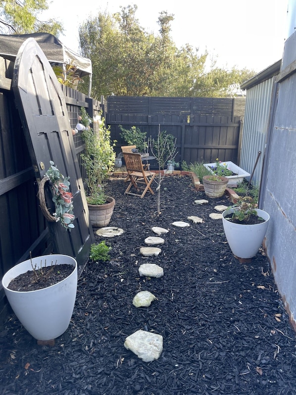 A garden pathway with stepping stones, surrounded by potted plants and a wooden chair.