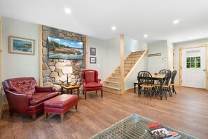A living room with a stone wall, a television, a fireplace, a red leather chair, a dining table with chairs, and a staircase. - Blackberry Woods | A Peaceful Maine Retreat in Hope (Hope)