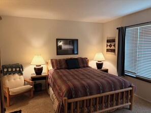 Ein Schlafzimmer mit einem Bett, zwei Nachttischen mit Lampen, einem Sessel, einem Wandspiegel und einem Fenster mit Jalousien.