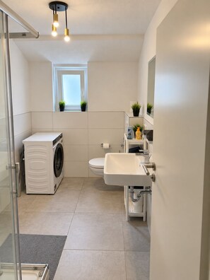 Ein modernes Badezimmer mit Waschmaschine, Toilette, Waschbecken und einem kleinen Fenster mit Pflanzen.
